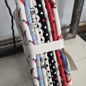 May include: A bundle of fabric squares tied with a white ribbon. The fabric features various patterns, including hearts, flowers, and arrows, in colors like red, black, and blue. The bundle is held together with a white ribbon.