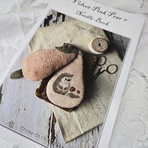 May include: Cross-stitch pattern for a pink pear with a hedgehog inside. The pattern is titled "Velvet Pink Pear + Needle Book" and is designed by Stacy Nash.