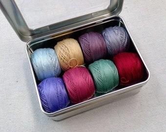 May include: A silver tin box with a clear lid, containing eight balls of yarn in various colors: light blue, beige, purple, gray, green, red, and two shades of purple.
