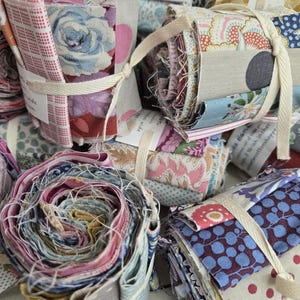 May include: A collection of fabric bundles, each tied with a cream-colored ribbon. The fabrics feature floral, polka dot, and geometric patterns in various colors, including pink, blue, and yellow. Some bundles are rolled, while others are stacked.