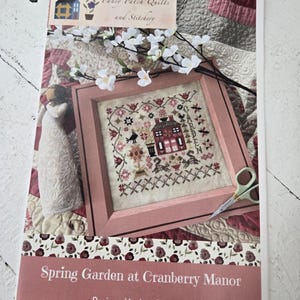 May include: A cross-stitch pattern titled "Spring Garden at Cranberry Manor" by Lori Pengelly. The design features a framed cross-stitch of a house and garden in a pink frame. The image includes scissors, a decorative figurine, and a floral arrangement.