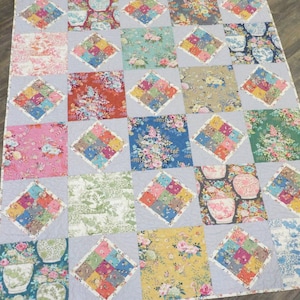 Può includere: Una trapunta con un design a patchwork che presenta vari motivi floreali e colori, tra cui rosa, blu, verde e giallo. La trapunta è composta da quadrati con motivi a forma di diamante all'interno di ogni quadrato.