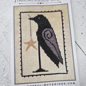 May include: Cross-stitch pattern showing a black crow with a gold star, with the text "Starring Russell Crow." The crow has a purple wing design. The pattern is framed with a dotted border and the website address www.artful-offerings.com.