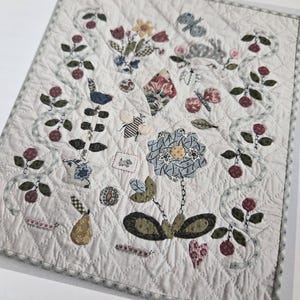 May include: A white quilt with a patchwork design featuring a variety of floral and animal motifs, including a butterfly, a bird, a bee, a pear, and a flower. The quilt is surrounded by a blue and white checkered border.