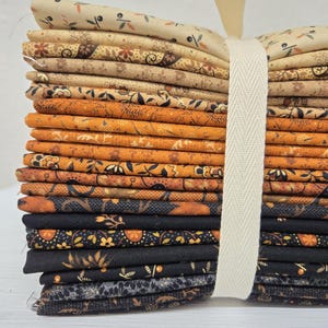 May include: A stack of folded fabric squares featuring various floral and geometric patterns in a color palette ranging from cream and tan to burnt orange, brown, and black. The fabric bundle is secured with a cream-colored herringbone ribbon.