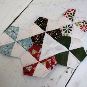 May include: A white fabric hexagon with various patterned fabric pieces sewn together. The fabric pieces are in shades of red, green, black, white, and blue. The fabric pieces are sewn together in a geometric pattern.