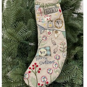 May include: A beige Christmas stocking with embroidered details, including the name "RORY", a clock, and the words "merry Christmas". The stocking is decorated with floral and festive motifs, and is displayed on a green Christmas tree.