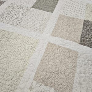 May include: A white quilt with a patchwork design featuring various shades of beige and gray. The quilt is made of different fabrics with subtle patterns, including small dots and floral motifs.