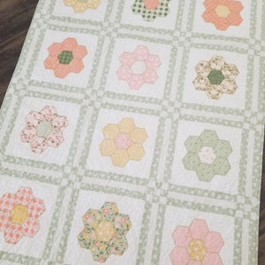 May include: A white quilt with a green border and a pattern of pink, yellow and green flowers. The flowers are arranged in a grid pattern.