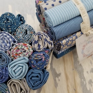 May include: A collection of blue and white fabric scraps rolled up and tied with a white ribbon. The ribbon has the text "Tilda Quilts" printed on it. The fabric scraps are in various patterns, including polka dots, stripes, and floral prints.