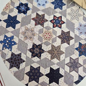 May include: A close-up of a circular quilt pattern featuring a repeating design of blue stars with various floral and geometric patterns. The quilt is made with a white background and has a grey and white geometric pattern in the background.
