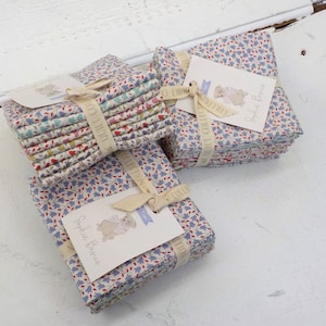 May include: A stack of fabric squares with a floral pattern in shades of blue, red, and white. The fabric is bundled with a white ribbon and a tag that says "Sophie B. Collection".