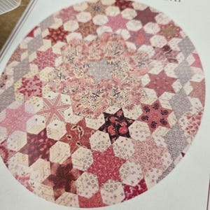 May include: A quilting pattern for a hexagon star-a-day quilt. The pattern features a circular design with a variety of pink, red, and white fabrics.