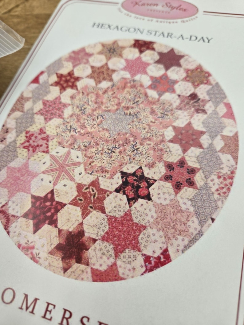 May include: A quilting pattern for a hexagon star-a-day quilt. The pattern features a circular design with a variety of pink, red, and white fabrics.