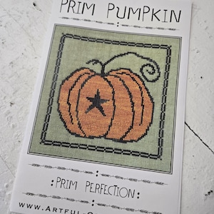 May include: A printed card featuring a cross-stitch design of an orange pumpkin with a black star on a green background. The text "PRIM PUMPKIN" and "PRIM PERFECTION" are displayed above and below the pumpkin image, respectively. The website address is also visible.