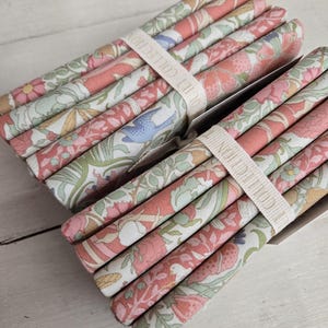 May include: Two bundles of floral fabric strips, each wrapped with a cream-colored band. The fabric features a pattern of pink, peach, green, and blue flowers and leaves. The band has the text "DUM COLLECTION" printed on it.