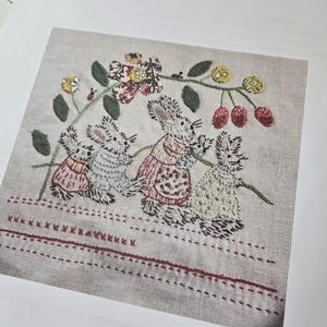 May include: A cross-stitch embroidery pattern featuring a group of three bunnies in pink and green dresses, surrounded by a vine with red berries and yellow flowers. The pattern is titled "Josephine".