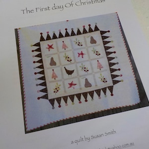 May include: A white quilt with a brown and red border. The quilt is made up of 16 squares, each with a different design. The designs include a pear, a Christmas tree, a star, a bird, and a holly sprig. The quilt is titled "The First Day of Christmas" and is by Susan Smith.