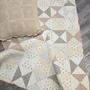 May include: A square beige crocheted pillow with a scalloped edge rests on a quilted blanket. The quilt features a patchwork design of triangles in cream, tan, and dark brown, with various small floral and geometric patterns printed on the fabric.