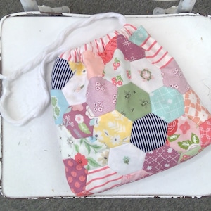 May include: A drawstring bag made of patchwork fabric with a hexagonal pattern. The fabric is a mix of floral prints, stripes, and solid colors in shades of pink, green, blue, and white.