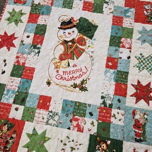 May include: A white patchwork quilt with a red, green, and blue color scheme. The quilt features a vintage-style illustration of a snowman with the text "A Merry Christmas!" in the center. The quilt is bordered with a red and white checkered fabric.