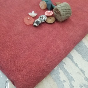 May include: A collection of assorted buttons in various colors and patterns, including pink, white, brown, and black, with a spool of brown thread on a red fabric background.