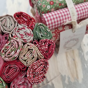 May include: A bundle of fabric rolls in red, green, and white patterns. The fabric rolls are wrapped in clear plastic and tied with a white ribbon. The ribbon has the text "Cotton + Tilda" printed on it.