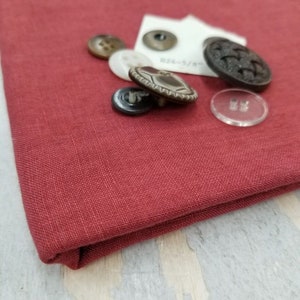May include: A close-up of a red fabric with a variety of buttons, including a silver button with a decorative design, a black button, a white button, and a brown button. The fabric has a subtle texture.