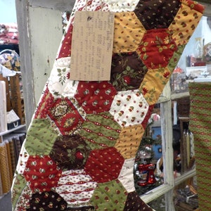 May include: A handmade Christmas stocking with a patchwork design. The stocking features a variety of fabrics in red, green, brown, and cream with floral and geometric patterns. A tag is attached with a button and twine.