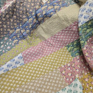 Op de afbeelding: Een patchwork quilt met verschillende bloempatronen in roze, blauw, groen, geel en grijs. De quilt is gemaakt van meerdere aan elkaar genaaide stofvierkanten, waardoor een textuuroppervlak ontstaat. Het ontwerp is een mix van kleine en grote bloemenprints.