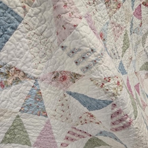 May include: Close-up of a quilted blanket with a patchwork design. The quilt features triangular patterns in various pastel colors, including pink, blue, and green, with floral and dotted prints. The quilt has a textured, quilted surface.