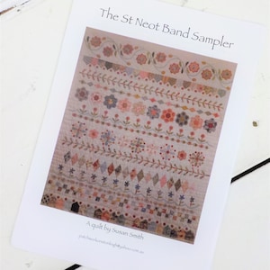 以下が含まれることがあります： 「The St Neot Band Sampler」というタイトルのキルトデザインが印刷された紙。キルトのデザインは、パステルカラーの花柄と幾何学模様の列が特徴です。「A quilt by Susan Smith」というテキストも表示されています。