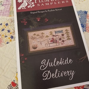 Peut inclure: Un modèle Plum Street Samplers intitulé "Yuletide Delivery" présentant un motif de point de croix. Le motif comprend un traîneau avec des rennes, des arbres et le texte "Dec 25". La couverture est sombre avec du texte blanc et une bordure décorative.