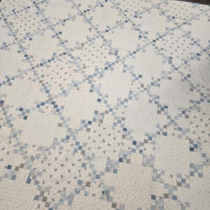 May include: A handmade quilt featuring a repeating pattern of light blue and gray squares on a white background. The quilt has a patchwork design with a diagonal layout, creating a textured appearance. The overall aesthetic is a blend of traditional and modern styles.
