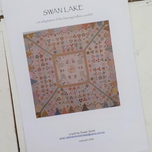 May include: A white paper with the title "SWAN LAKE" in black text. The text below the title reads "(an adaptation of the Dancing Dollies coverlet)". The paper features a photo of a quilt with a variety of colours and patterns. The text at the bottom of the paper reads "a quilt by Susan Smith", "email: patchworkonistonleigh@yahoo.com.au", and "copyright 2009".