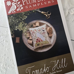 May include: A white card with a brown border and the text "Plum Street Samplers" in red and black. The card features a photo of a small, hand-stitched sampler with a red and green tomato design and the text "Tomato Hill" in cursive. The text "Original Designs by Paulette Stewart" is also included.