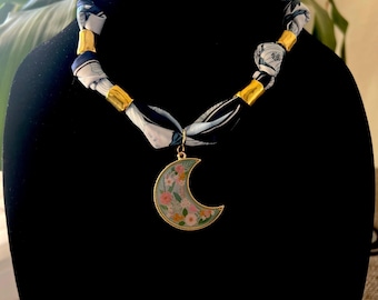 May include: A necklace featuring a silk scarf with a black and white floral pattern. The necklace has gold-colored metal accents and a crescent moon pendant with a floral design in pink, yellow, and green.