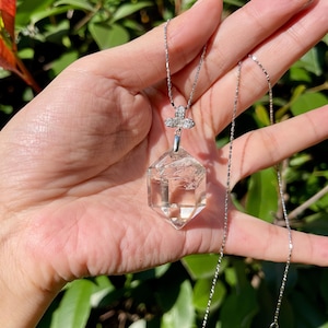 以下が含まれることがあります： 透明な水晶ペンダントネックレス、シルバーチェーン付き。ペンダントは多面的な六角形です。ネックレスは、透明な石が付いた小さな装飾的な蝶の留め金が特徴です。