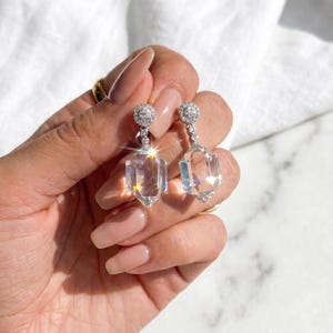 Op de afbeelding: Een paar heldere, facetgeslepen kristallen oorbellen met een geometrische vorm. Elke oorbel heeft een kleine, ronde, met diamanten bezette bovenkant en een hangend, langwerpig zeshoekig kristal. De oorbellen worden door een hand omhoog gehouden.