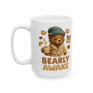 May include: White ceramic mug featuring a cartoon bear in a robe and beanie, holding a coffee cup. The mug has the text "BEARLY AWAKE" in brown letters. The design includes coffee beans, a croissant, and a small alarm clock.