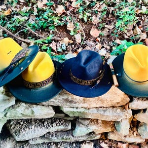 May include: Four fedora hats with a dark brim and a yellow crown are displayed on a stone wall. One hat is navy blue, and the others have a yellow crown. Each hat has a decorative band and feather accents.