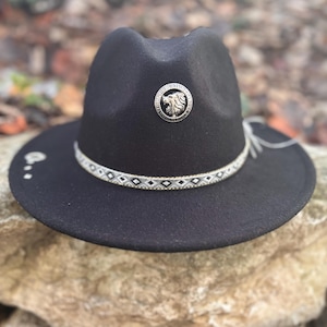 Peut inclure: Un chapeau fedora noir avec une bande décorative argentée à motifs géométriques. Le chapeau a un emblème de lion argenté à l'avant et une ficelle décorative sur le côté. Le chapeau repose sur une pierre.