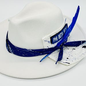 May include: A white fedora hat featuring a blue bandana wrapped around the crown. A rectangular patch with the text PHI BETA SIGMA is attached to the side, accompanied by a white fabric piece with black paint splatters and a blue feather.