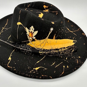 Op de afbeelding: Een zwarte fedora-hoed met gouden verfspatten over de kroon en de rand. De hoed is versierd met een goudkleurige broche met een kroon en vleugels met een barnsteenkleurige steen, een gouden ketting en twee veren, &eacute;&eacute;n geel en &eacute;&eacute;n zwart.