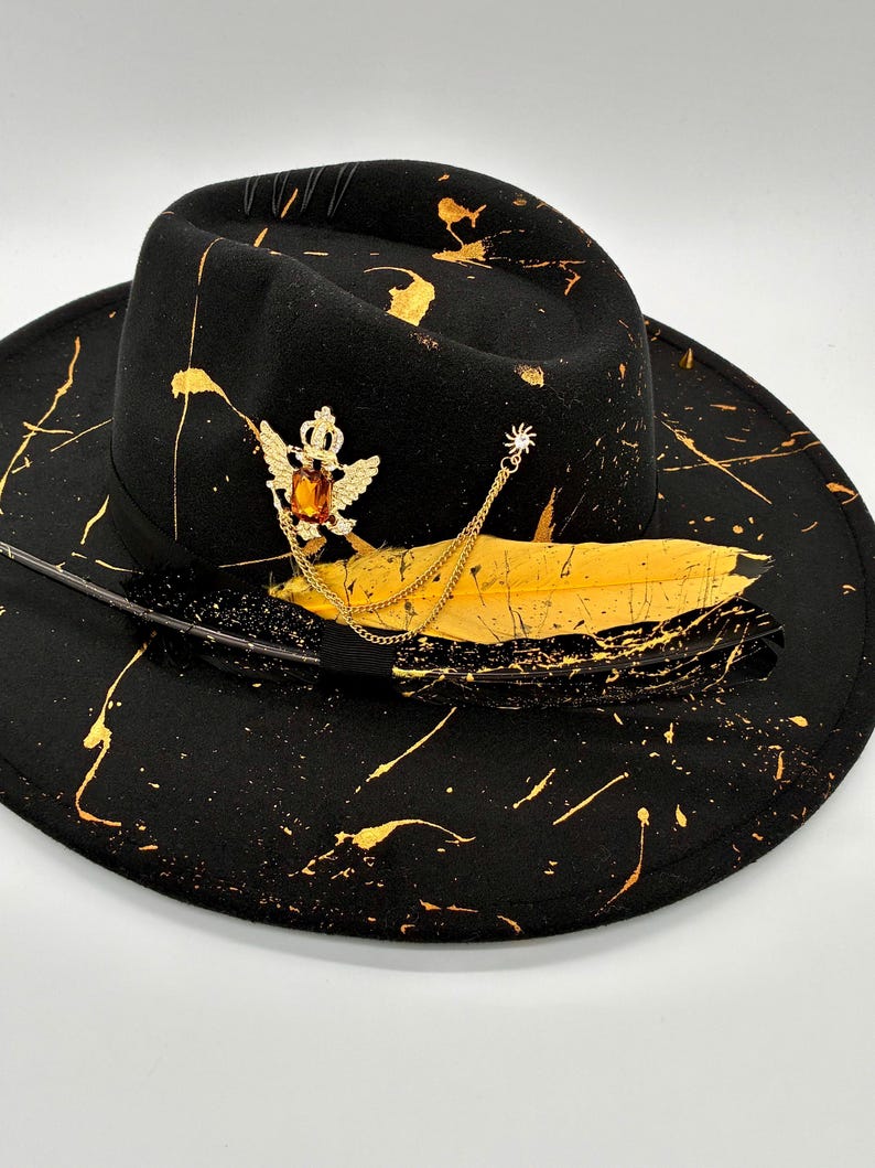 Op de afbeelding: Een zwarte fedora-hoed met gouden verfspatten over de kroon en de rand. De hoed is versierd met een goudkleurige broche met een kroon en vleugels met een barnsteenkleurige steen, een gouden ketting en twee veren, &eacute;&eacute;n geel en &eacute;&eacute;n zwart.