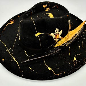Op de afbeelding: Een zwarte fedora hoed met een brede rand en een zwarte lintband. De hoed is versierd met gouden verfspatten, een goudkleurige metalen broche met een centrale barnsteenkleurige steen en een lange, gele en zwarte veer.
