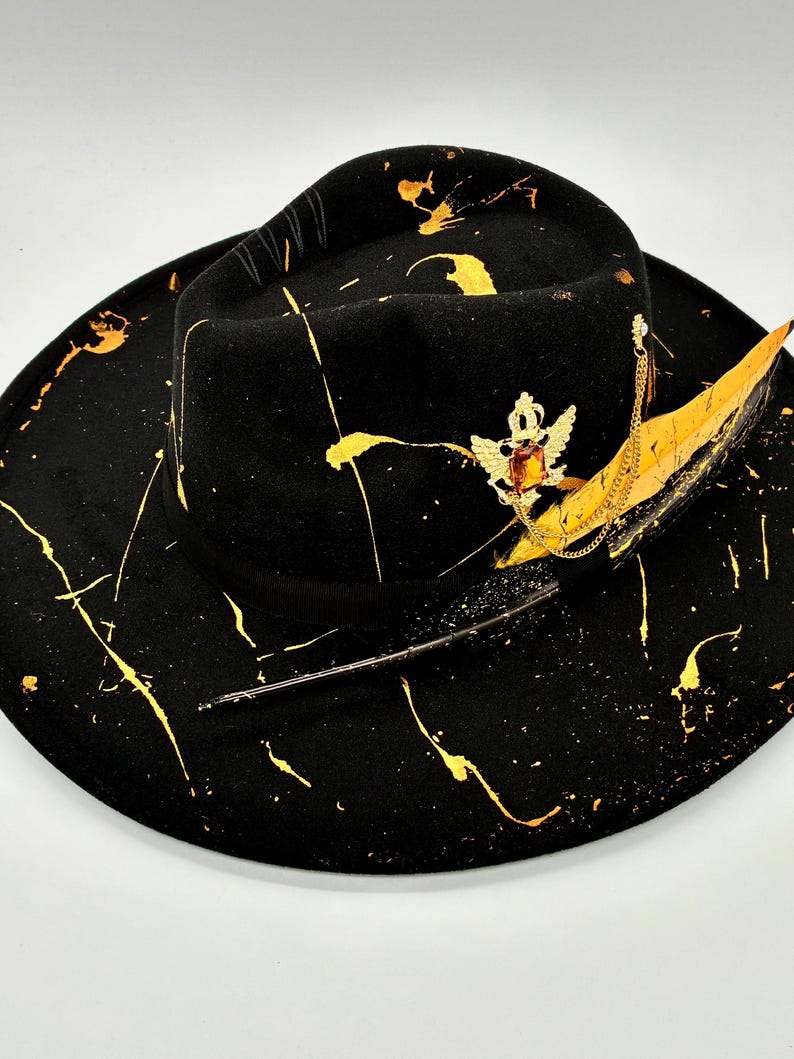 Op de afbeelding: Een zwarte fedora hoed met een brede rand en een zwarte lintband. De hoed is versierd met gouden verfspatten, een goudkleurige metalen broche met een centrale barnsteenkleurige steen en een lange, gele en zwarte veer.