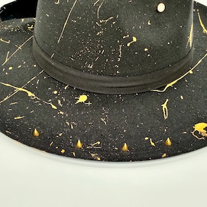 Op de afbeelding: Een zwarte fedora hoed met een goudkleurig verfspatpatroon over de kroon en de brede rand. De hoed is voorzien van een zwarte grosgrain lintband rond de basis, een enkele gouden stud op de kroon en een rij kleine gouden kegelvormige studs.