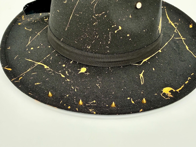 Op de afbeelding: Een zwarte fedora hoed met een goudkleurig verfspatpatroon over de kroon en de brede rand. De hoed is voorzien van een zwarte grosgrain lintband rond de basis, een enkele gouden stud op de kroon en een rij kleine gouden kegelvormige studs.