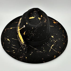 Op de afbeelding: Een zwarte fedora hoed met een brede rand en een ingedeukte kroon, versierd met gouden verfspatten over het oppervlak. Een zwarte grosgrain lintband omcirkelt de basis van de kroon, geaccentueerd door een goudkleurige veer en metalen details.